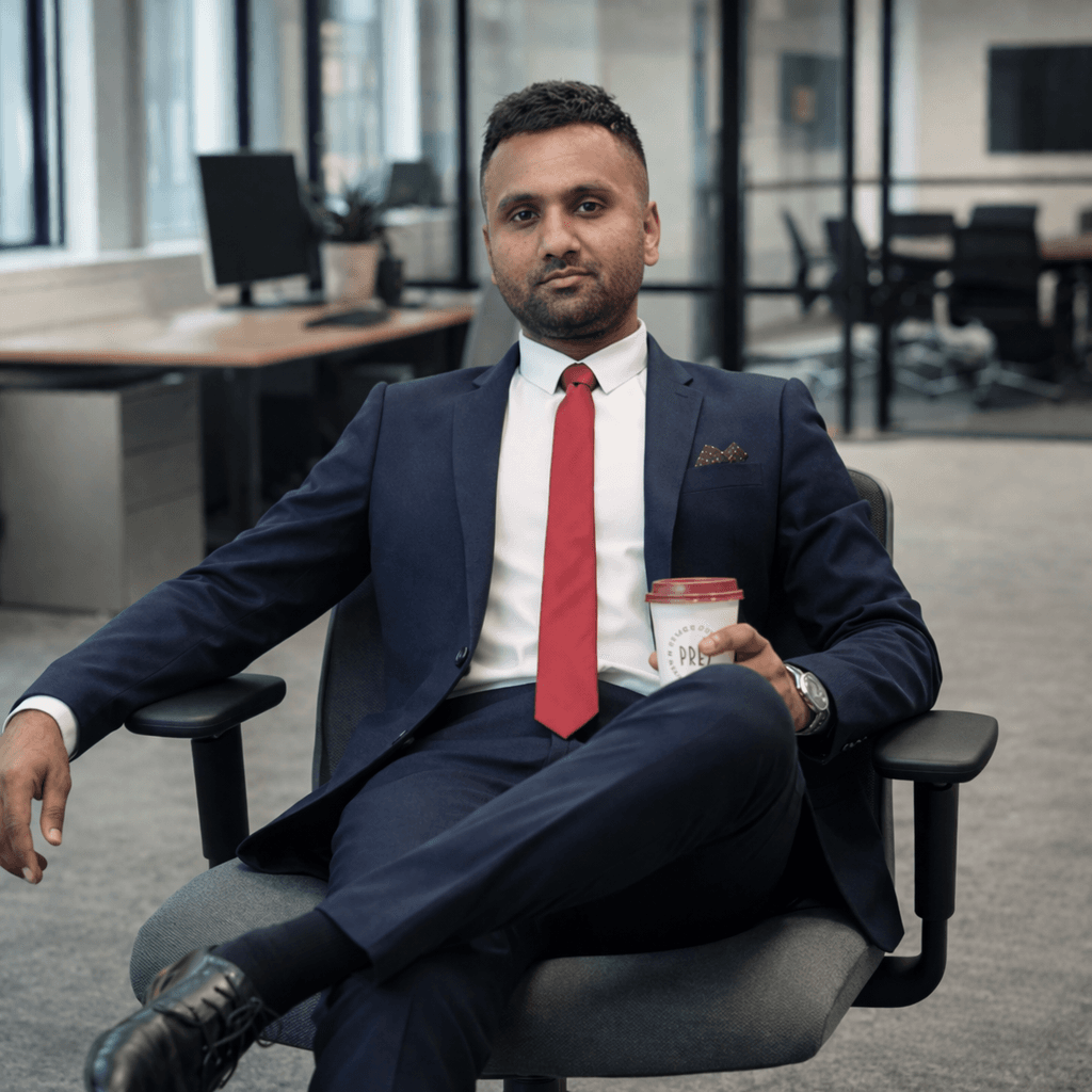 Man in navy suit and red tie sitting in office chair holding coffee cup.